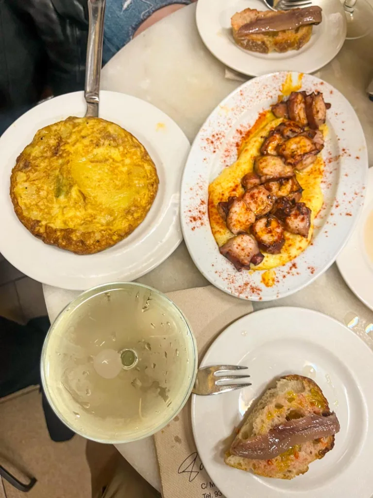 an overview shot of a dinner table at el xampanyet in barcelona, with a full spanish tortilla, octopus a la gallega, toast with anchovies and a cava glass