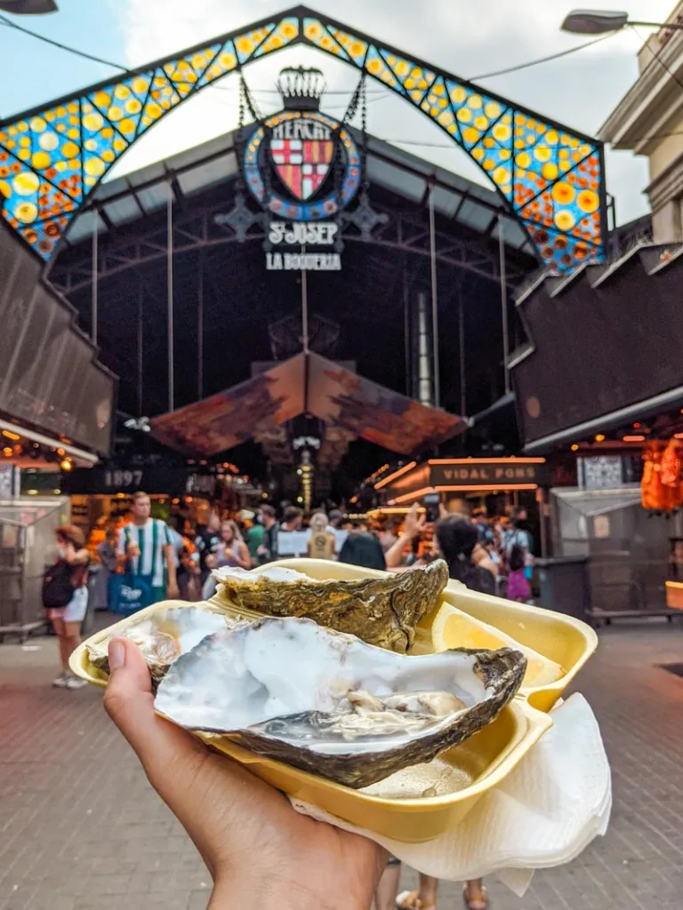 hand holding a paper plate with oysters, with the view of la boqueria market  in barcelona at the back