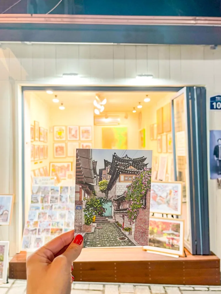 hand holding a postcard with a painting of the buckhon hanok village in front of de art little shop in seoul