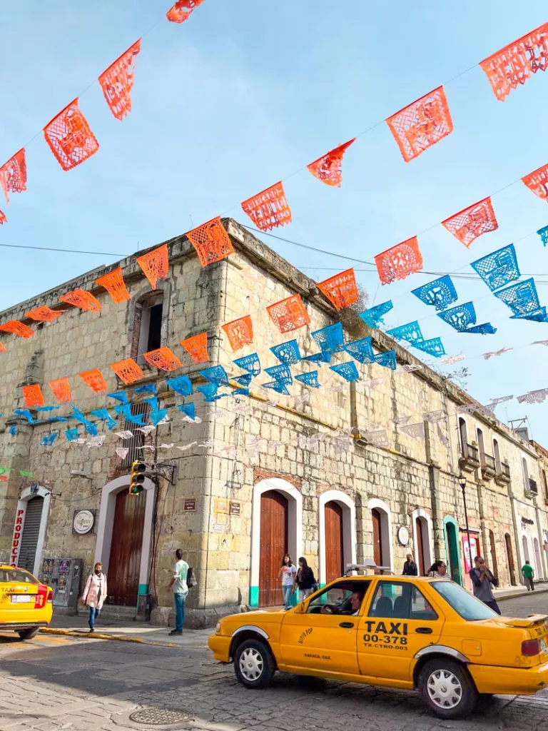 Colonial buildings, and papel picado dangling over a street in in Oaxaca City as a taxi drives past