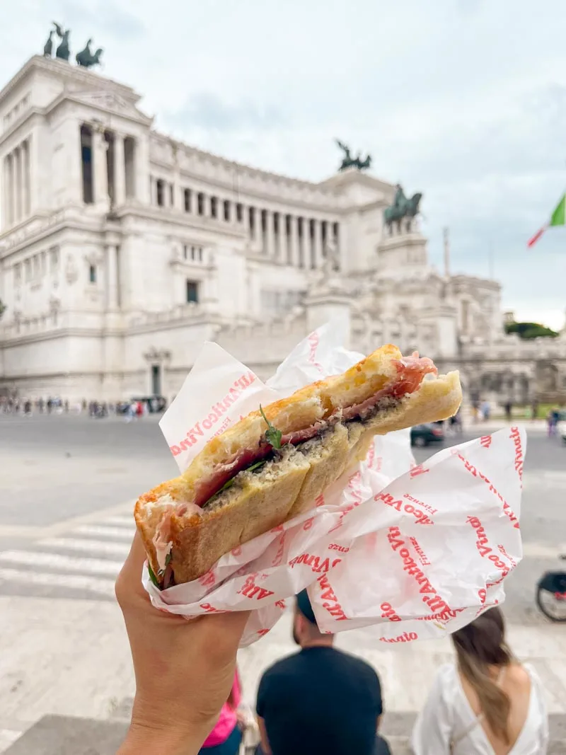 hand holding a panino from all antico vinaio in front of the vittoriano monument in rome