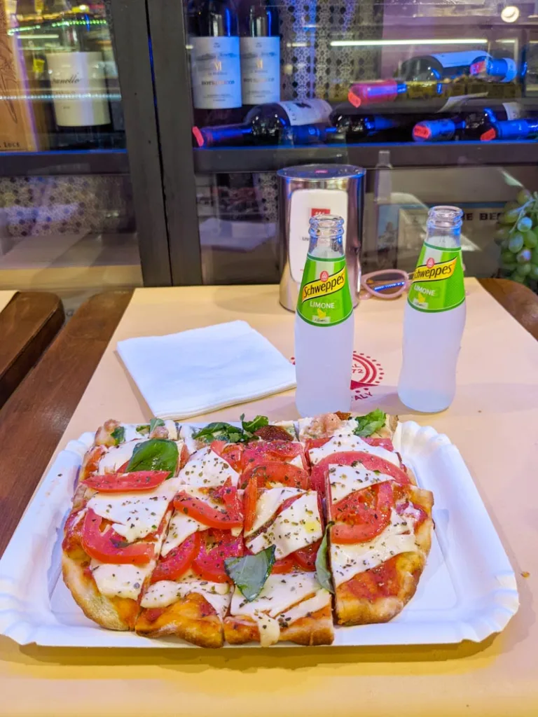 cut up pizza pieces on a paper plate and two schweppes glass lemonades at a table at roscioli bakery
