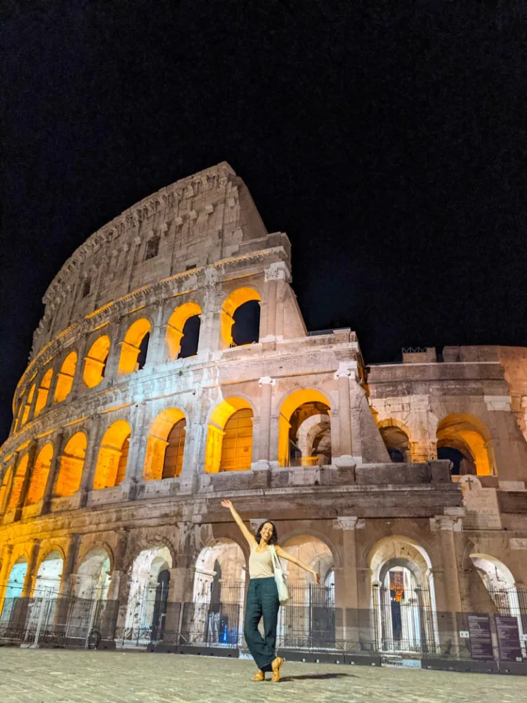 woman standing with arms open wide in front of an empty colosseum at night in rome