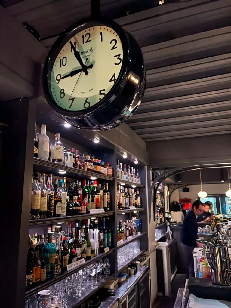 a train station clock, and shelves stacked with liquor bottles behind the bar at a cocktail bar in ebisu, tokyo