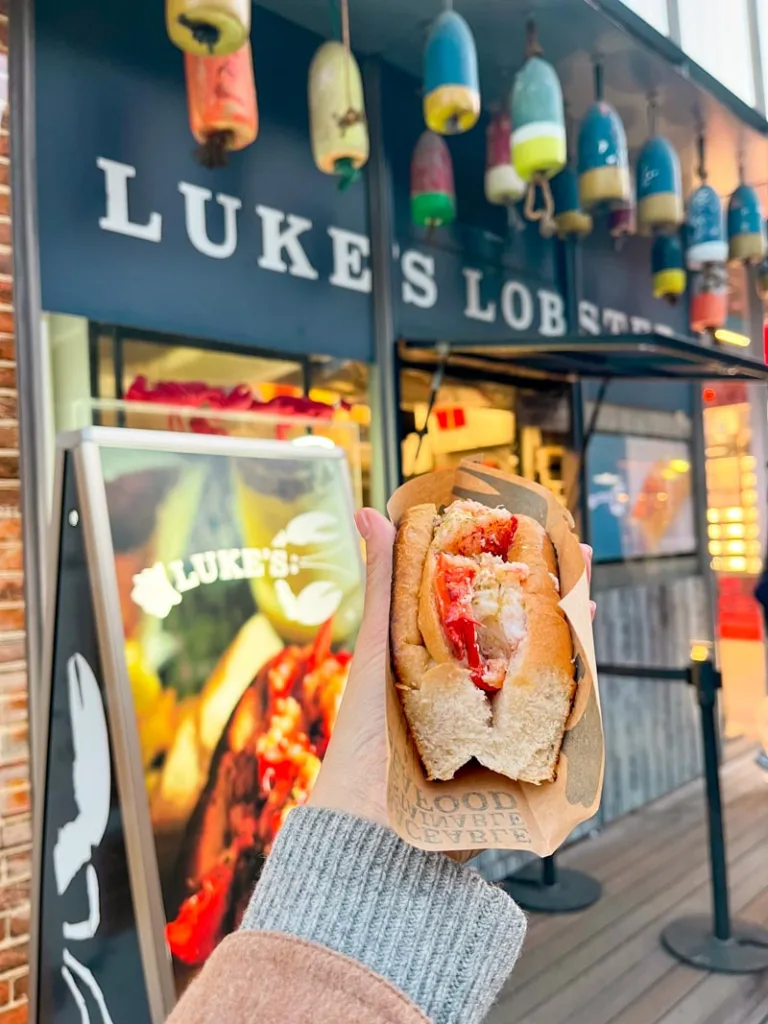 hand holding a lobster roll with a sign of luke's lobster small food stand behind, in shibuya, tokyo