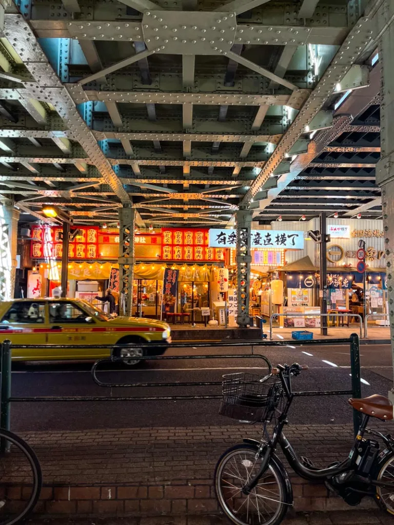 view of the warm glowing light of a restaurant across the street, under a bridge, and a taxi driving by in shimbashi, tokyo