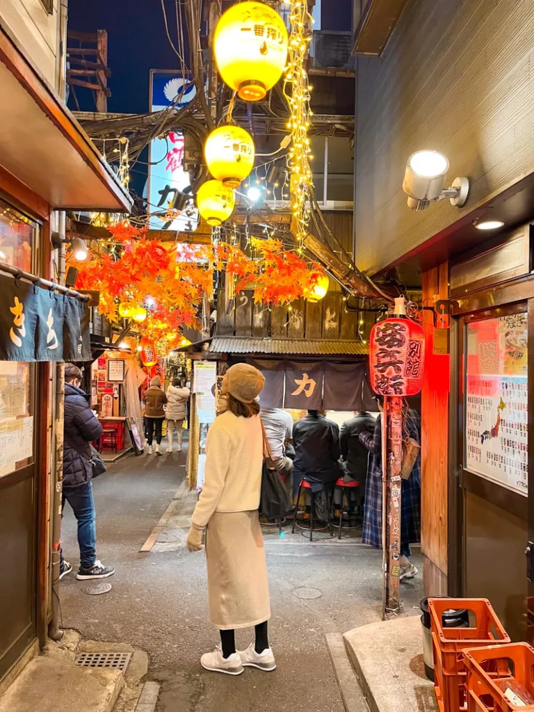 woman standing in the middle of a snug alley lined with restuarants at the famous shinjuku omoide yokocho in tokyo