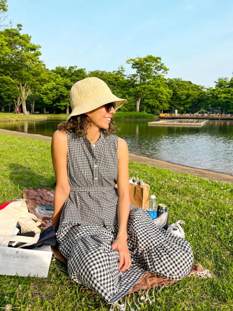 woman in a black and white checkered dress, wearing a small sun hat and sunglasses sitting on the grass at yoyogi park in tokyo