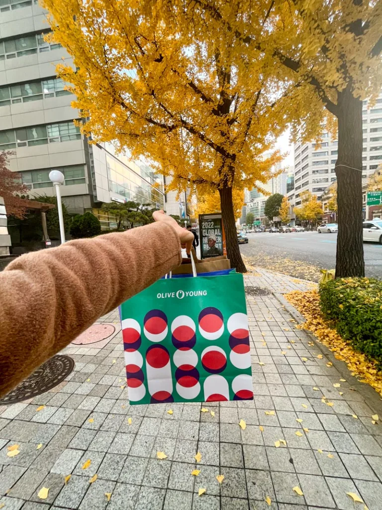 hand holding an olive young shopping bag in the middle of a street with a view of ginkgo trees in seoul