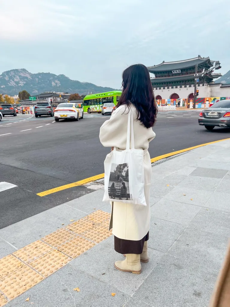 stylish korean woman waiting for a green light, wearing a cream colored coat, brown pants, beige suede boots and a tote bag in seoul