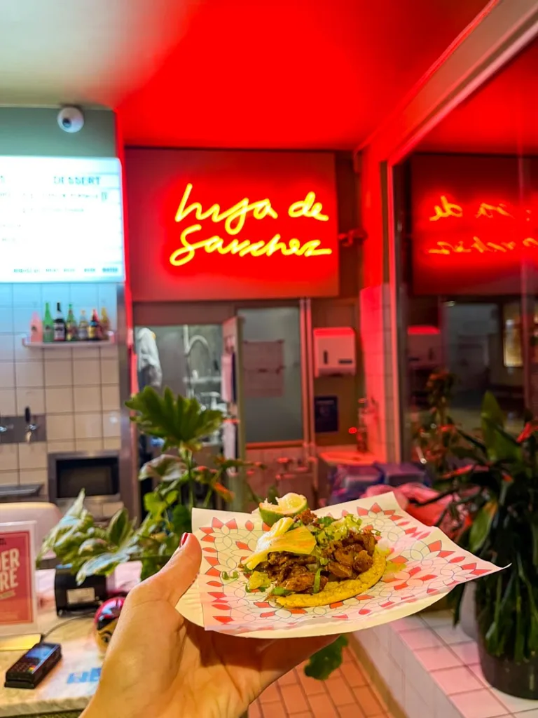 hand holding a plate of tacos with a red neon sign reading "hija de sanchez" behind, in copenhagen