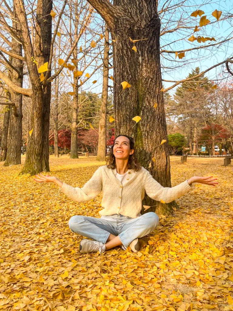woman sitting on the floor throwing yellow ginkgo leaves in the air while the floor is covered with them at nami island, south korea