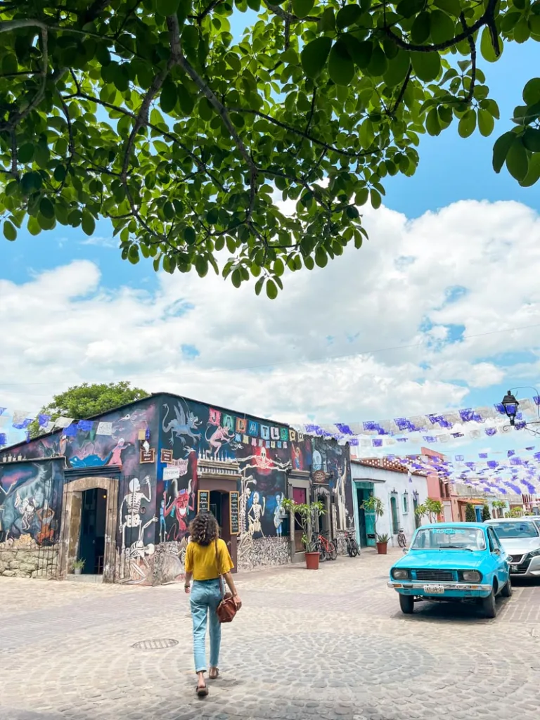 woman walking towards a mural-covered cafe with colorful papel picado covering the street next to it in jalatlalco, oaxaca city