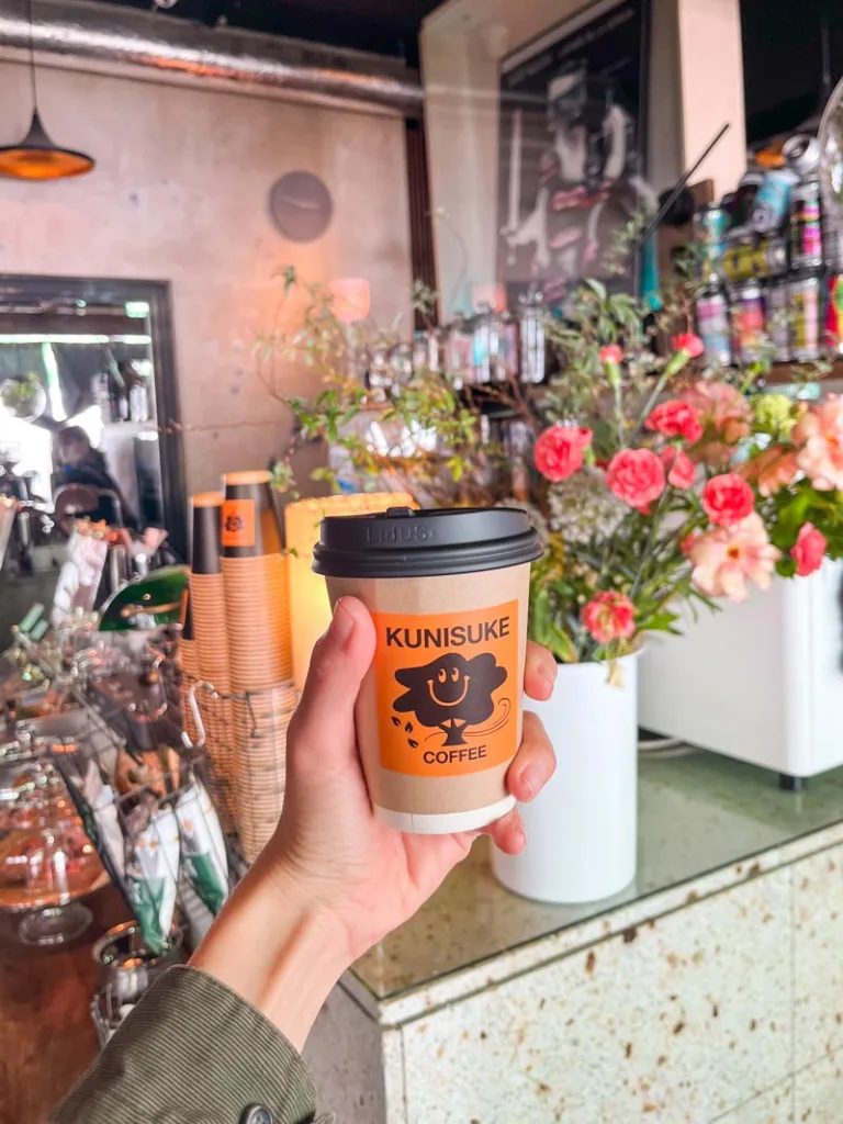 hand holding a to-go coffee cup with a sticker of kunikuse coffee shop in tokyo, flowers on display at the coffee bar behind