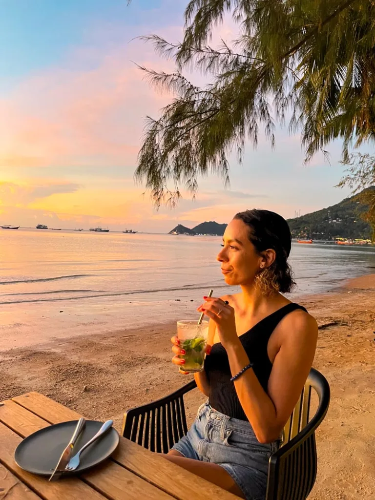 woman holding a drink sitting on a chair, looking out at the ocean on golden hour in koh tao, thailand