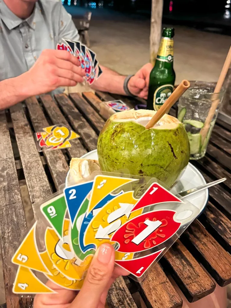 two people sitting across from each other holding uno cards, with a beer, a cocktail, and a coconut in the center of the table at a bar in koh tao, thailand