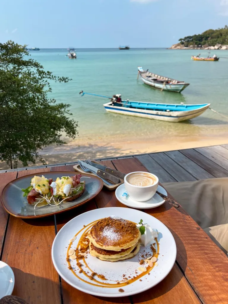 a plate of pancakes, eggs benedict and a coffee on a wooden table at koppee espresso bar with a direct view of the ocean in koh tao thailand