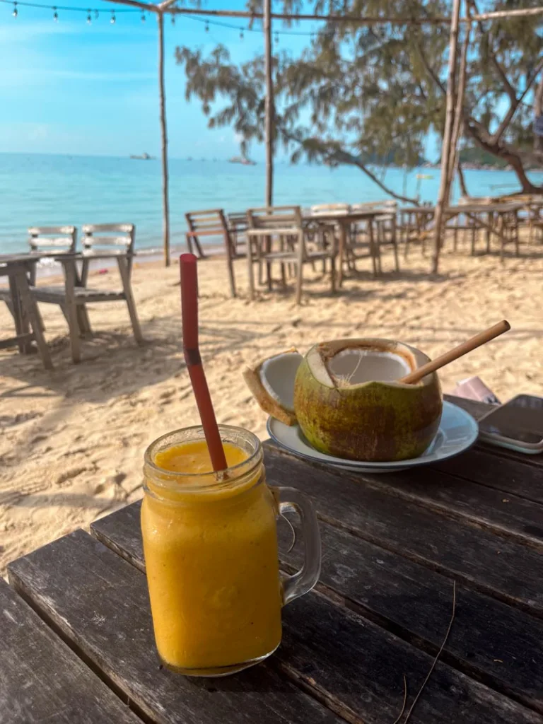 an open coconut on a plate with a straw, and a mango smoothie on a wooden table on the sand at sairee beach in koh tao, thailand