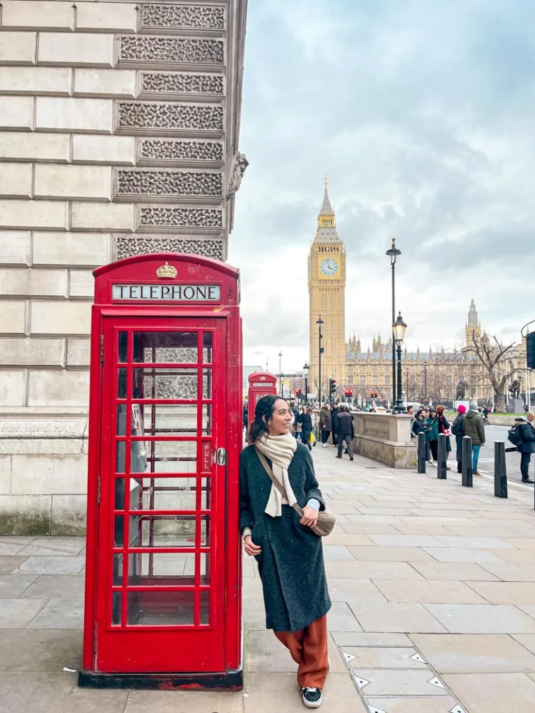 woman leaning against a red phone booth in london with a view of big ben behind