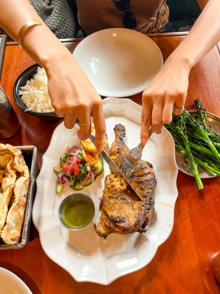 two hands holding a fork and knife cutting into half a roasted chicken, a side of rice, and side of broccolis, on a table at dishoom restaurant