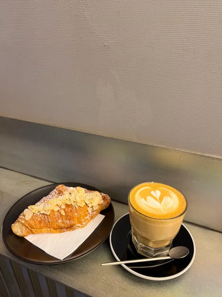 a latte in a transparent glass and an almond croissant on a metal bar at lift coffee shop in notting hill, london