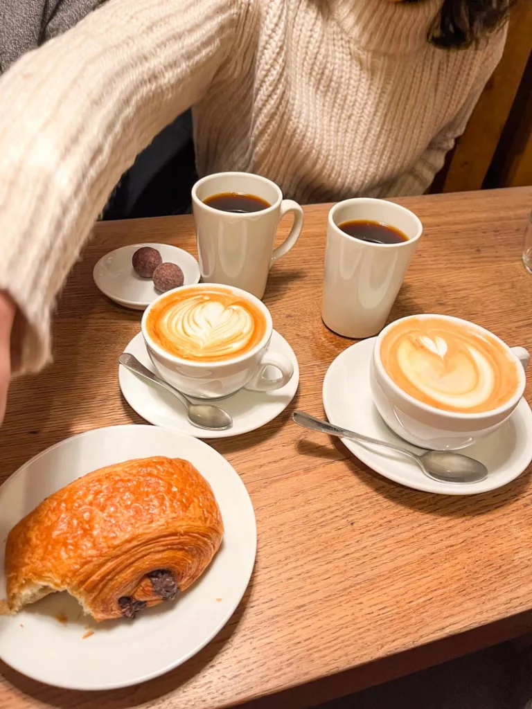 two lattes, an arm reaching over for a bitten pain au chocolat and two black coffees on a wooden table at monmouth coffee in london