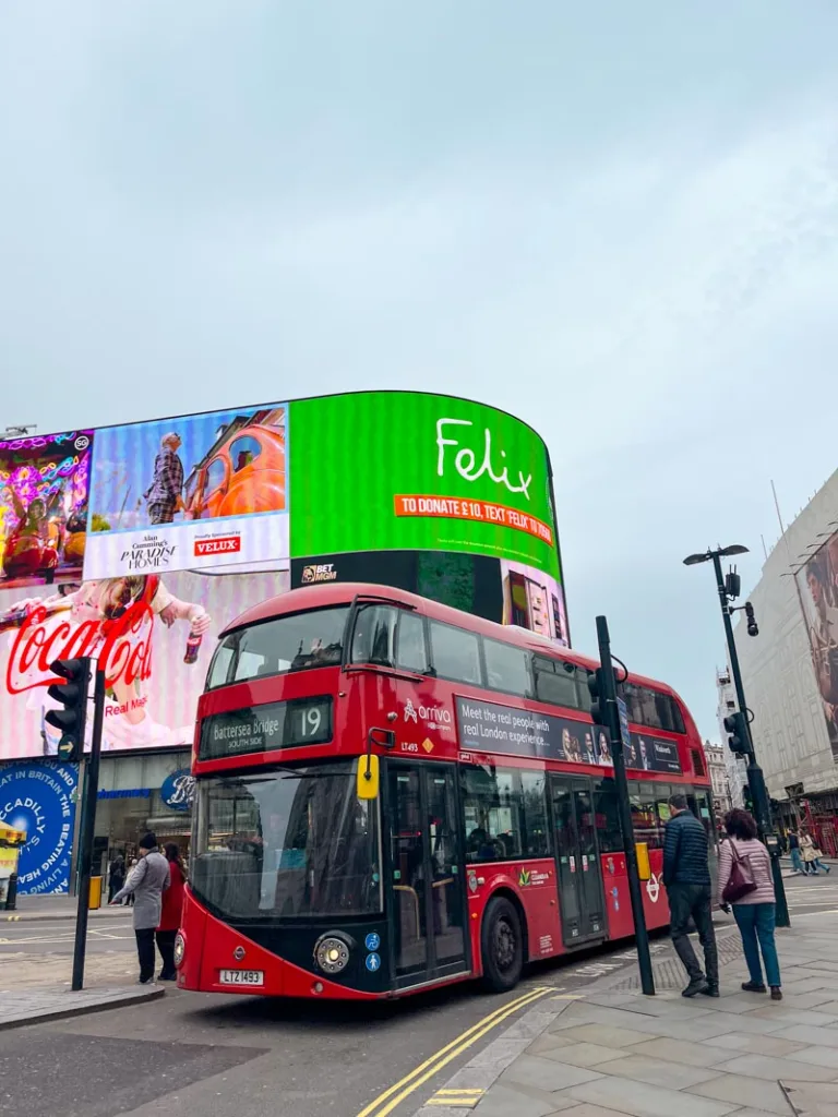 red double decker bus driving through piccadilly circus in london with a view of a big LED screen behind