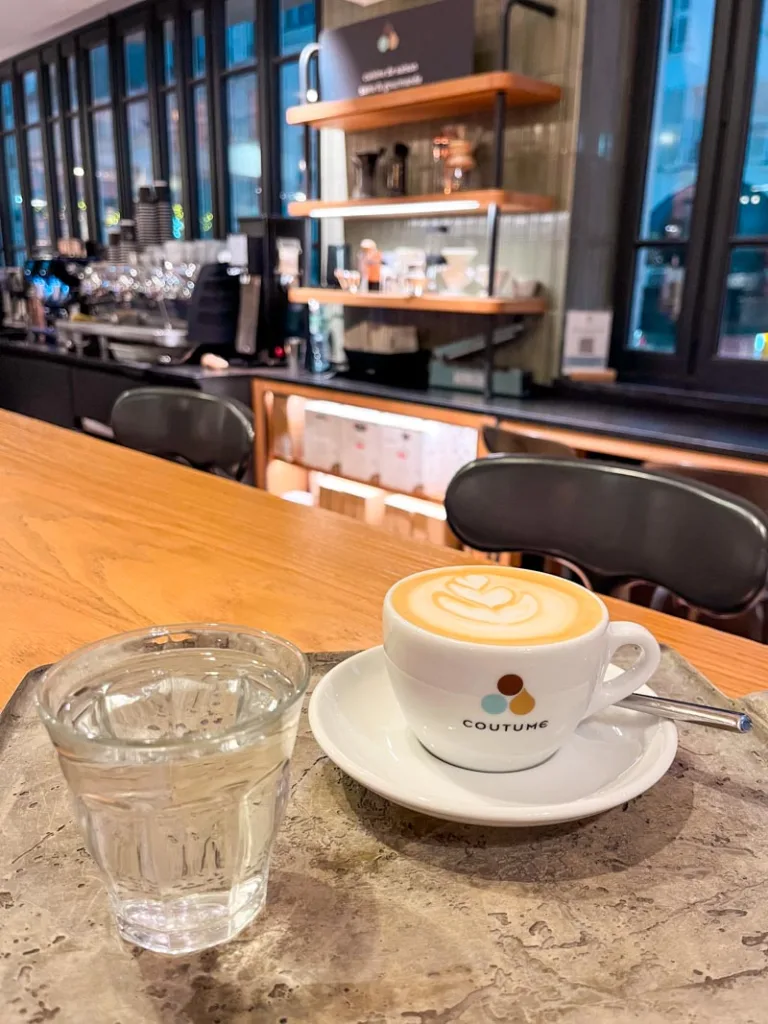 a small capuccino in a white cup, a small glass of water, on a silver tray, on a wooden bar at coutume specialty coffee shop in paris