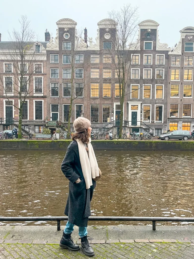 woman standing next to a canal in amsterdam with cute dutch houses at the back