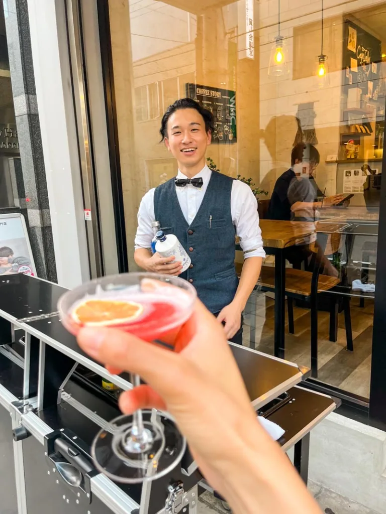 Hand holding a cocktail outside of Cafe Tales in Osaka, with the bartender of Bar Kadom smiling in the back