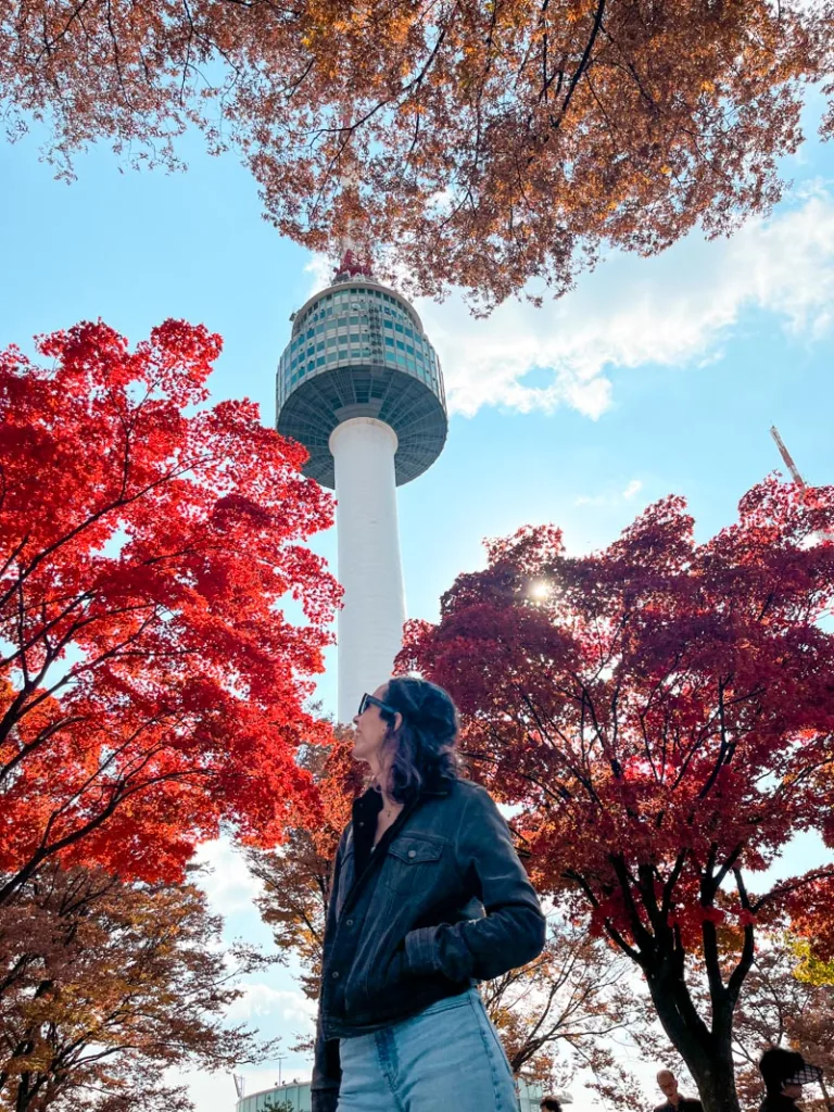 Woman standing in front of the N-Seoul tower framed by trees with bright red leaves
