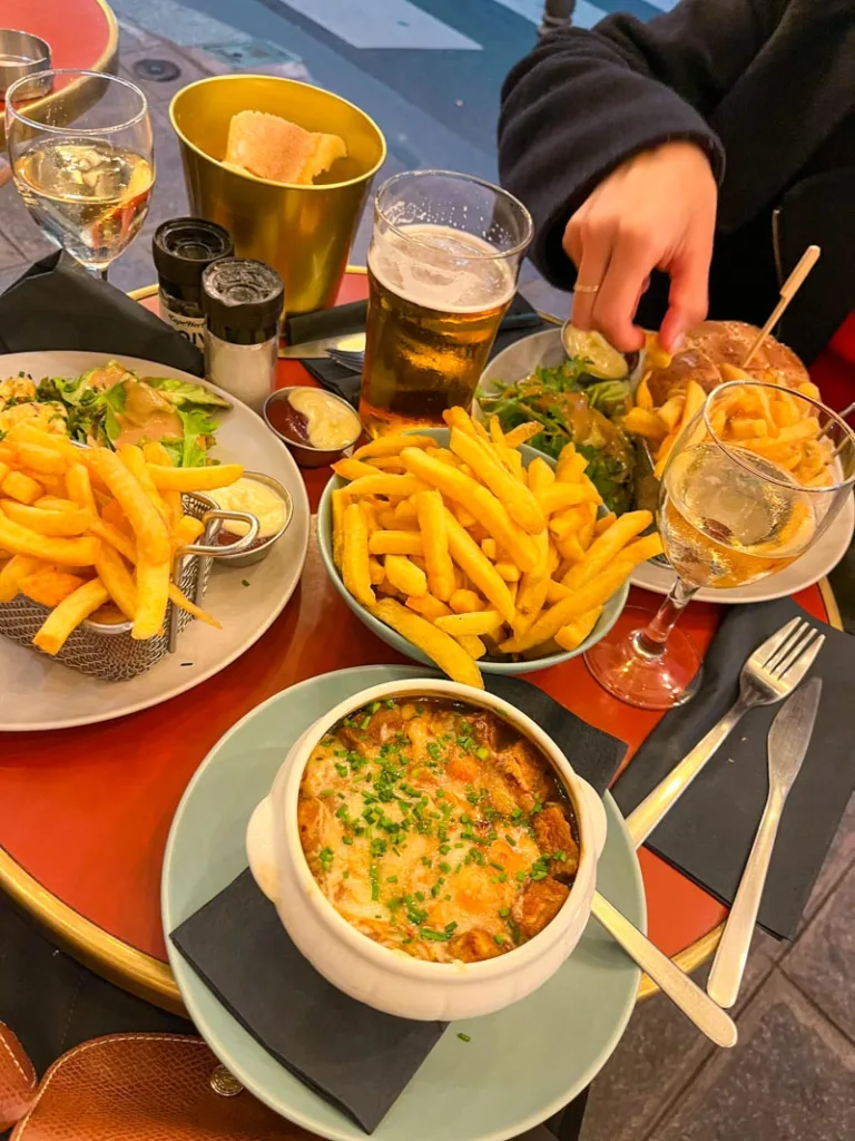 small french cafe table packed with plates, an onion soup, french fries, a burger, an omelette, a bread basket, two champagne glasses and a beer at le balto in paris