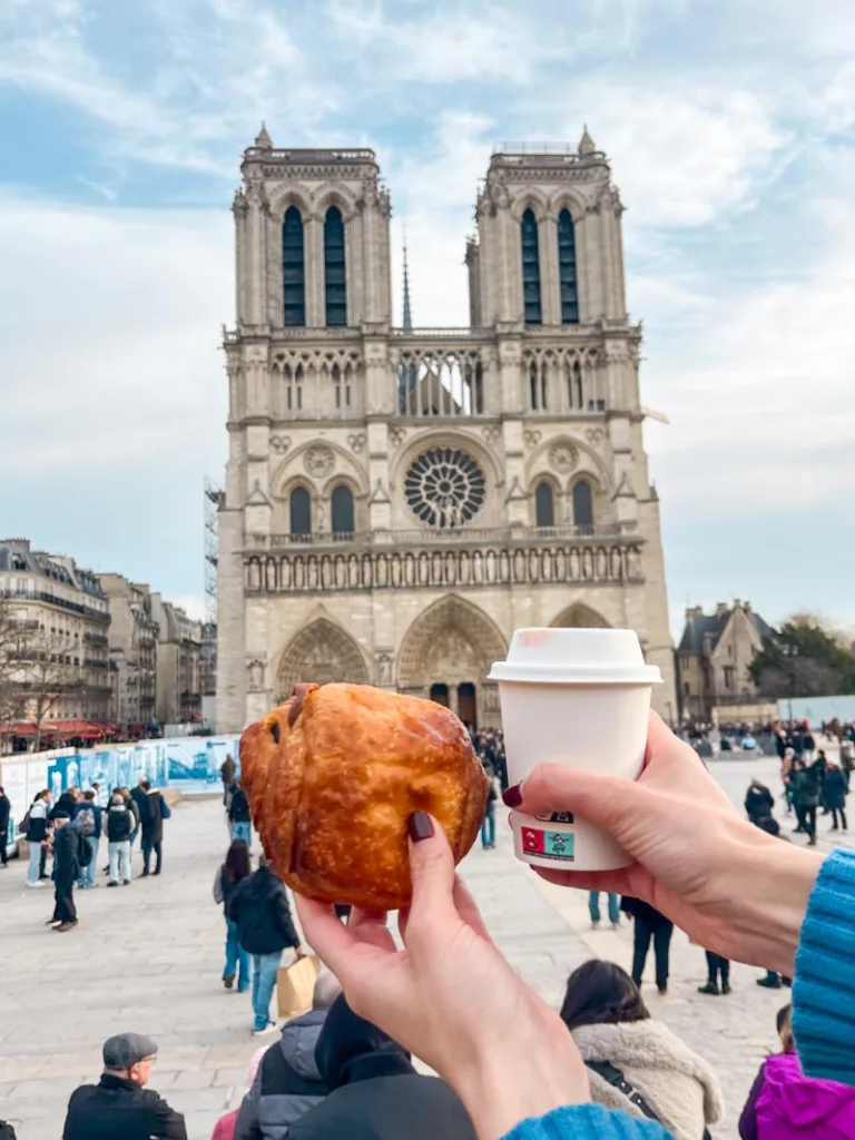 two hands holding a pain au chocolate and a coffee in front of the notre dame cathedral in paris