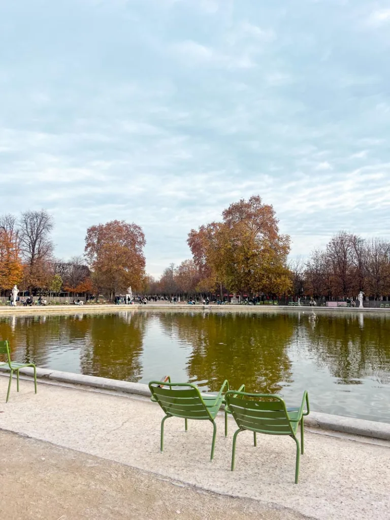 Two green chairs by the pond in jardin des tuileries in paris, with autumnal-colored trees in the back