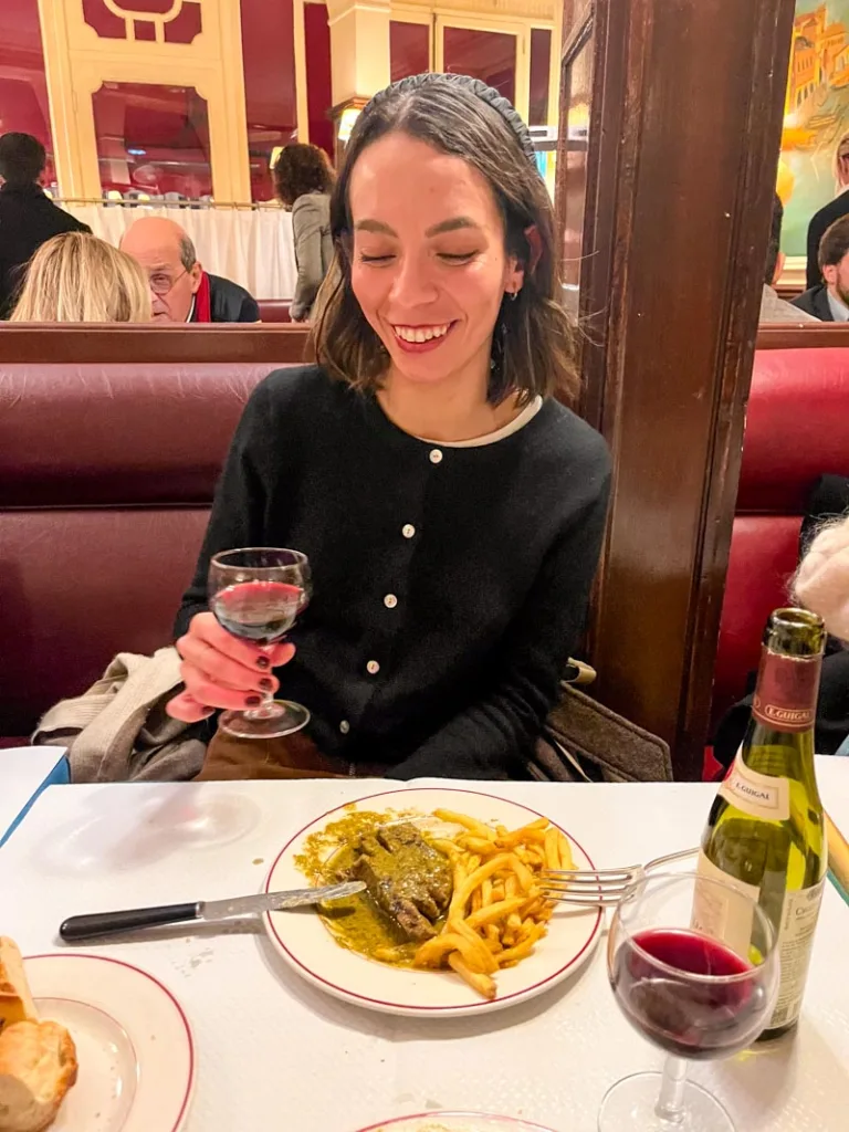 woman smiling, holding a glass of red wine, and a plate of steak frites in front of her at relais de l'entrecote in paris