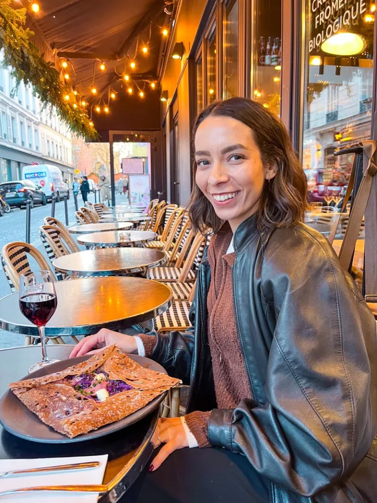 woman smiling with a plate with a crepe and a glass of red wine on an outdoor cafe table at cafe breizh in paris