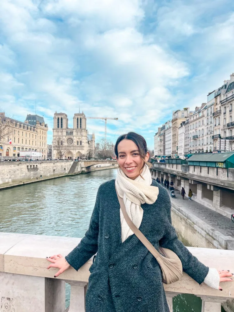 Woman smiling, standing on a bridge with a view of Notre Dame Cathedral in the back in Paris