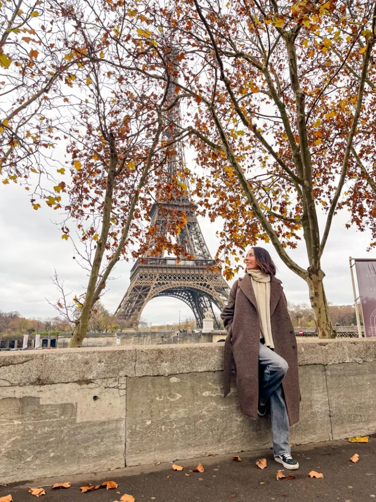 Woman smiling with a view of the Eiffel tower in the back peaking through the autumn leaves on the trees in Paris