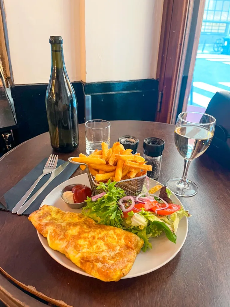 plate with an omelette, side salad, a fries portion, a glass of white wine next to it and a glass water bottle at le balto bistro in paris