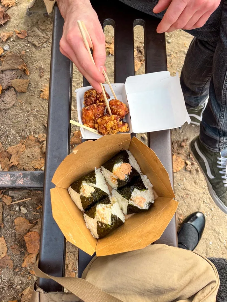 box of four onigiris and a small box with fried chicken pieces from omosubi gonbei on park bench in paris