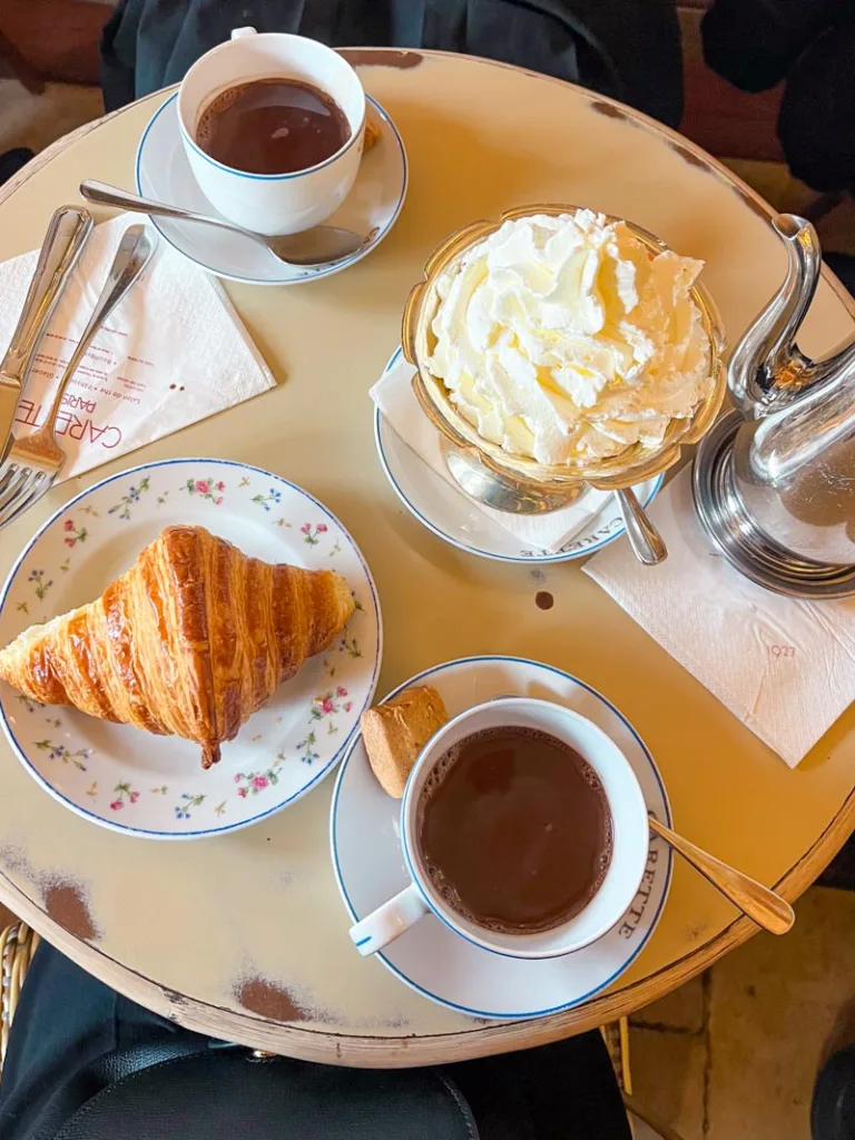 small french cafe table with two hot chocolate cups, a croissant and a serving cup with a mountain of whipped cream at carette in le marais