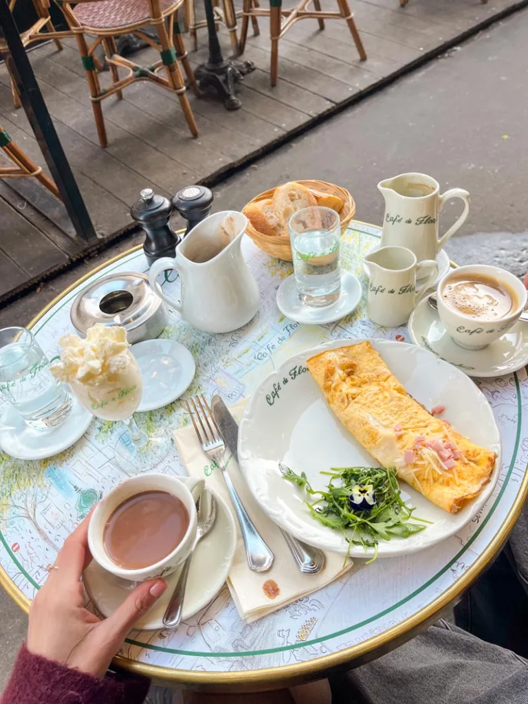 spread on a table at cafe de flore: a ham and cheese omelet, black coffee, a hot chocolate with a wine glass with whipped cream next to it, a bread basket and a water jug