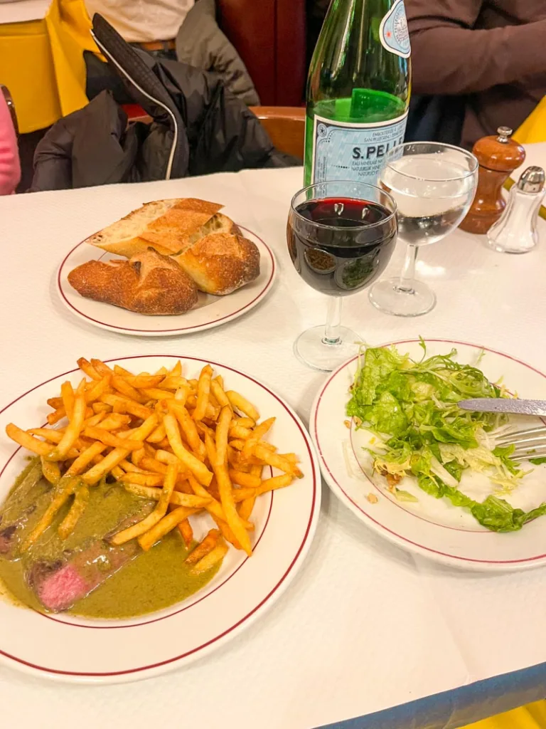 plate of steak frites, a side salad, a bread plate and a glass of wine at a table at relais de l'entrecote