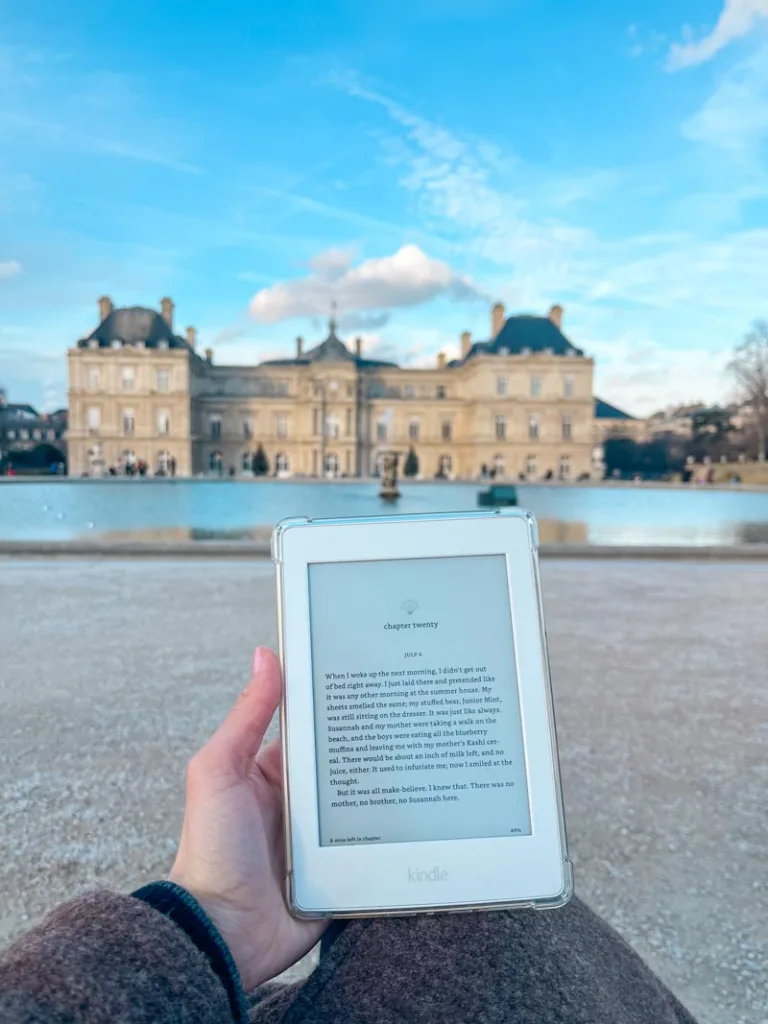 hand holding a kindle with a view of the pond and the palace of luxembourg at the back at the jardin du luxembourg, paris