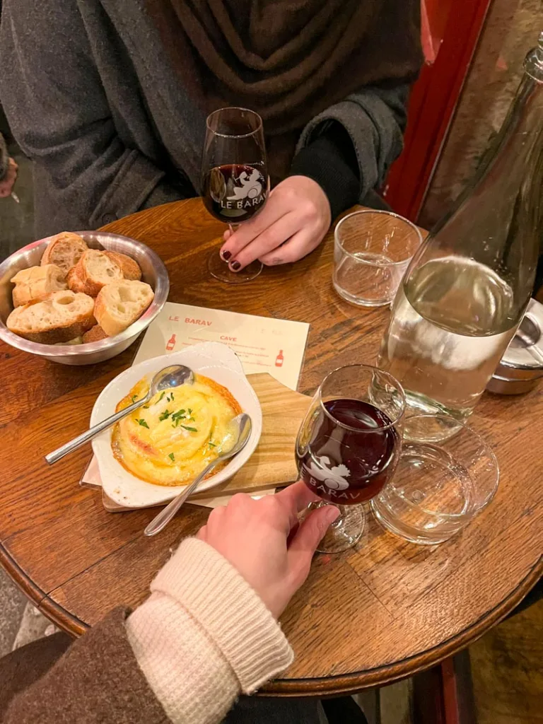 two people sitting at a table with two glasses of red wine infront and a small bowl of melted cheese at le barav wine bar in paris