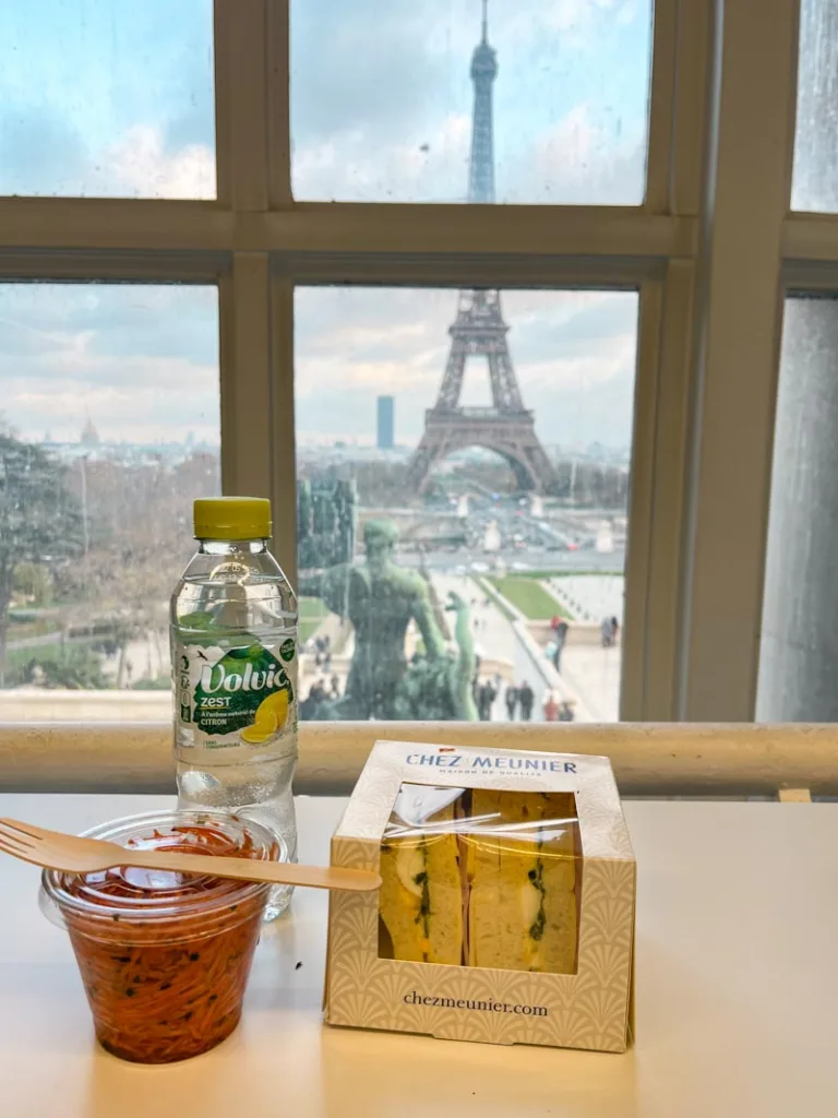 a packed sandwiched, a carrot salad and a water bottle on a window table at the library of contemporary art in paris with a view of the eiffel tower