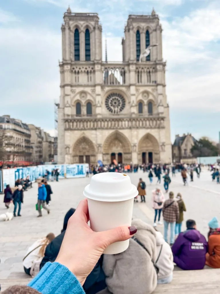 hand holding up a coffee cup to-go with a view of notre dame cathedral in the back