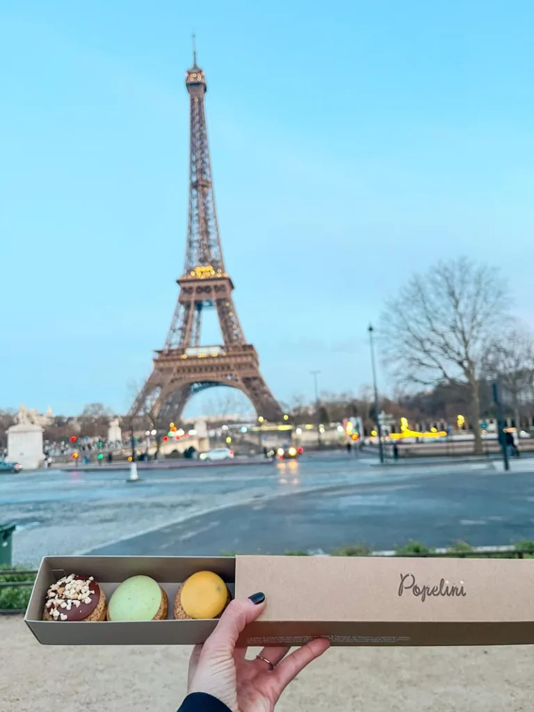 hand holding a box of popelini's cream puffs with a view of the eiffel tower behind