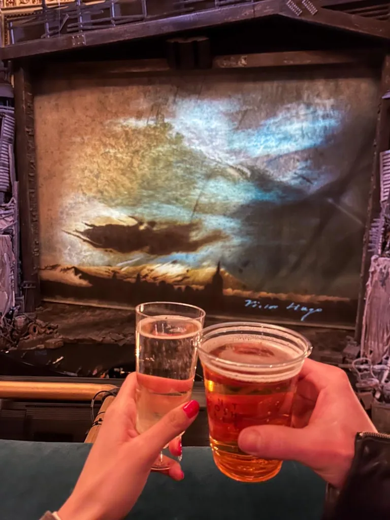 two hands cheering drinks in front of Les miserables musical curtain inside the theatre