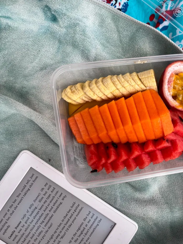 kindle and a plastic tupperware with cut up fruit on a beach blanket in koh tao